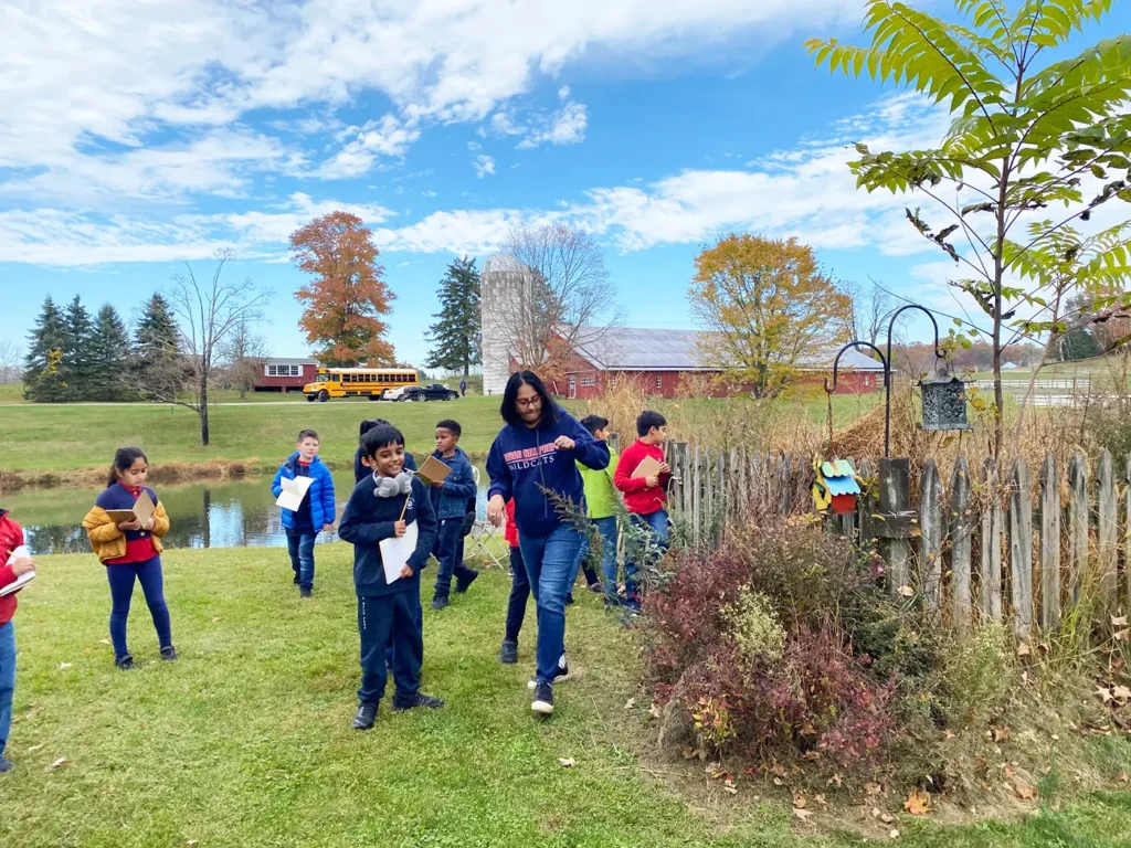 Elementary students on an outdoor field trip near a farm, exploring nature with journals beside a red barn