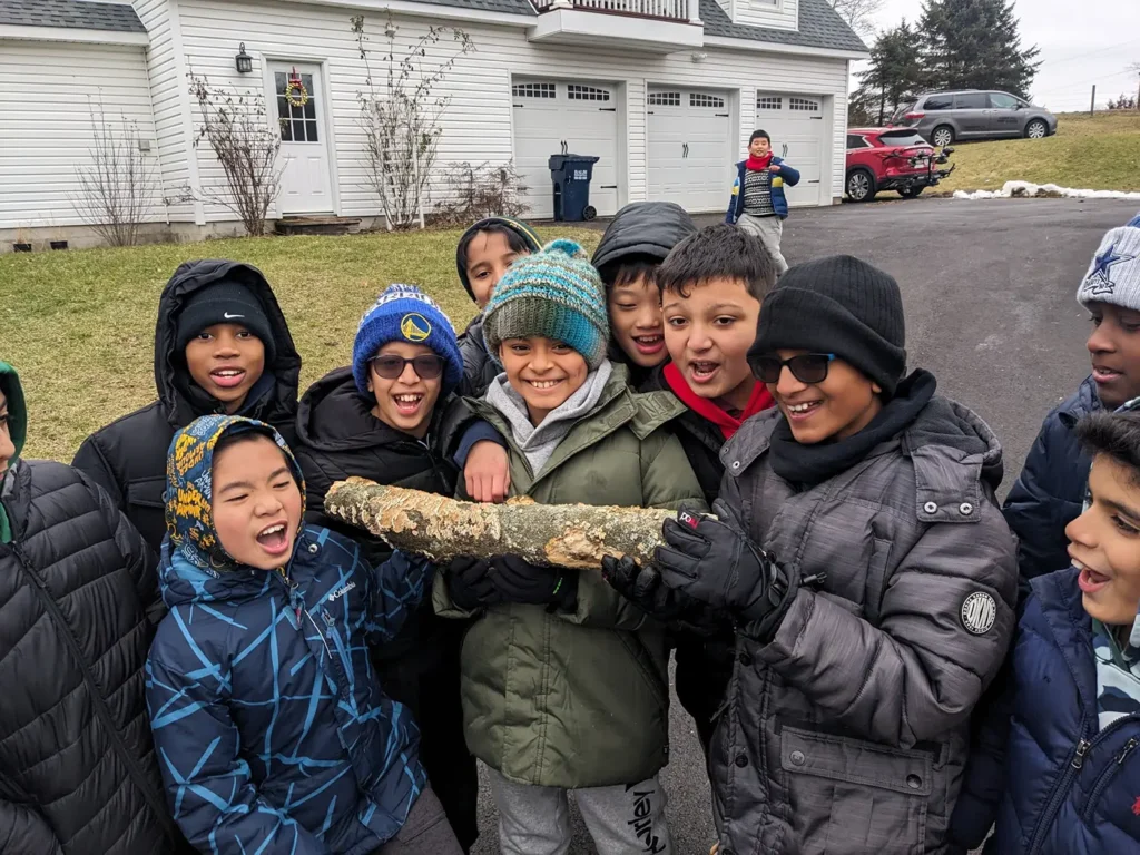 Group of bundled-up boys smiling and holding a log outside a house during a winter outdoor activity
